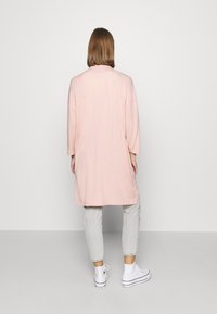 Cárdigan rosa de gran tamaño con mangas largas y un corte holgado, combinado con jeans grises y zapatillas blancas. Textura suave, diseño minimalista.