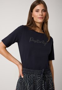 Marineblaues T-Shirt mit kurzen Ärmeln, das mit "Positivity!" in silbernen Nieten verziert ist. Kombiniert mit einem gemusterten Rock mit elastischem Bund in dunklen Farbtönen.