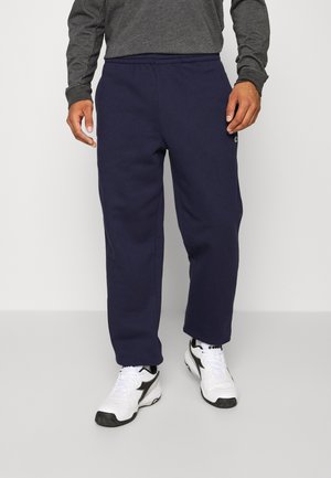Personne portant un pantalon de survêtement bleu marine, un haut à manches longues gris et des baskets blanches, debout devant un fond clair uni.
