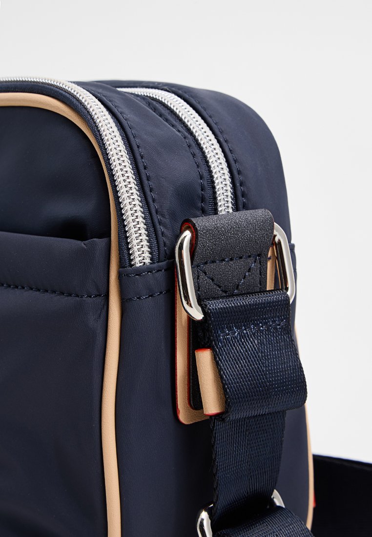 Sac en nylon bleu marine avec double zip argenté, accents beiges et une sangle texturée. Présente des accessoires en métal et un design rectangulaire compact.