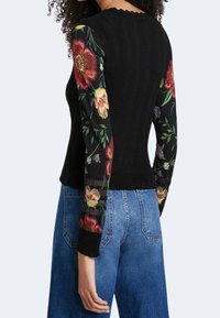 Musta ribbineulottu neule, jossa on kukkaommeluksin koristellut verkko-hihoja, joissa on kirkkaita punaisia ja keltaisia kukkia tummalla taustalla.