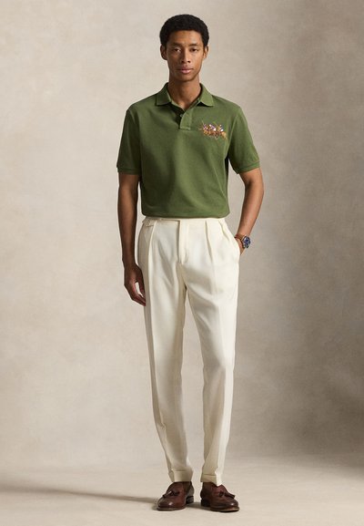 Άνδρας που φοράει ελιάς πράσινο πόλο με κεντήματα ιππέων, άσπρο πλισέ παντελόνι, καφέ μοκασίνια με φούντες και ρολόι χειρός, στέκεται μπροστά σε ουδέτερο φόντο.
