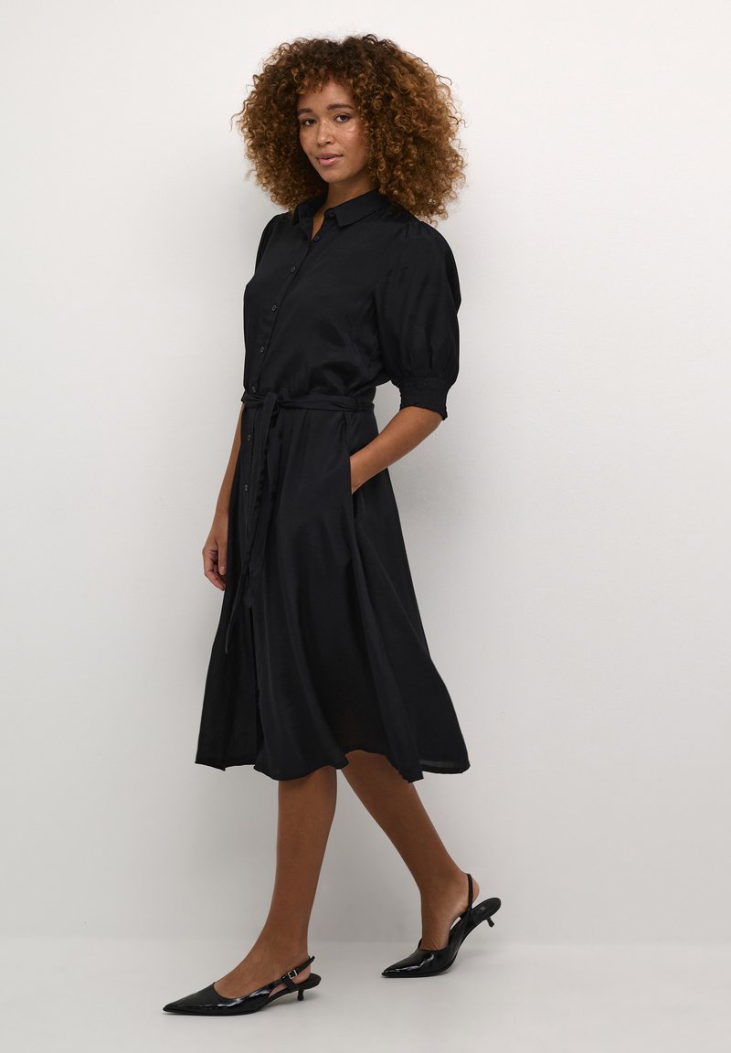 Robe noire en tissu lisse, avec un devant boutonné, des manches bouffantes, une ceinture à nouer à la taille et une jupe fluide.