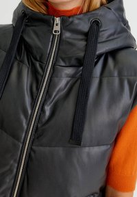 Veste sans manches matelassée en cuir noir avec un col montant, une fermeture éclair argentée et des cordons texturés. Dotée d'une surface matelassée et d'un design rembourré.