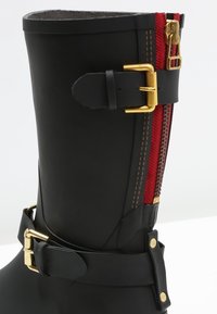 Black rubber boot with red zipper detail, gold hardware, and a black adjustable buckle strap. Features stitching accents along the seam.