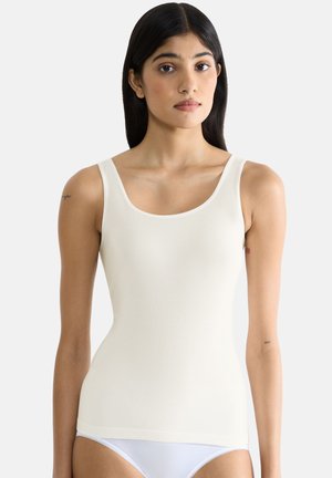 Junge Frau mit langen schwarzen Haaren, die ein off-white ärmelloses Oberteil und weiße Unterwäsche trägt, steht vor einem schlichten hellen Hintergrund.