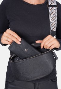 Black leather crossbody bag with a textured surface, oval shape, silver zipper, and a patterned strap. A matching wallet is being retrieved from inside.