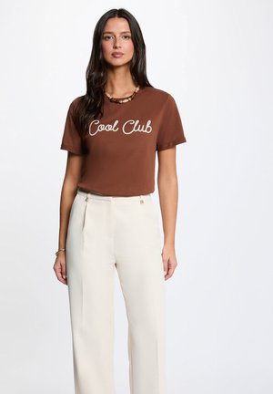 Kvinde med langt mørkt hår iført en brun "Cool Club" t-shirt, hvide bukser med høj talje og vide ben, perlekæde og armbånd på ensfarvet baggrund.