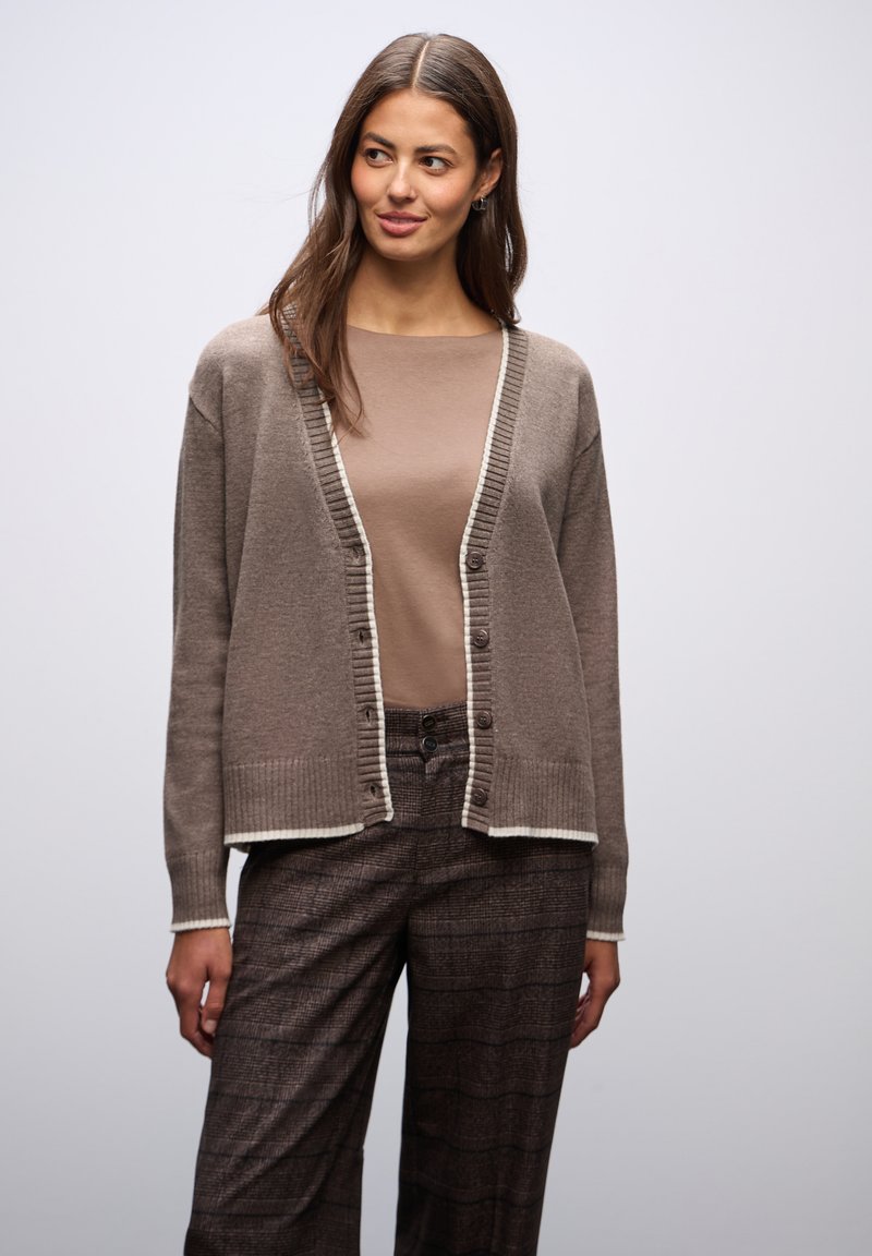 Cárdigan marrón con bordes acanalados y detalles en crema, de frente abierto, combinado con una blusa lisa beige y pantalones de pierna ancha marrones de cuadros.