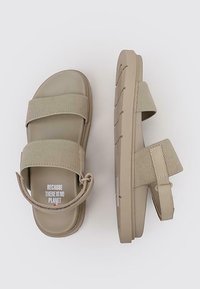 Sandalias beige con dos tiras de banda texturizadas, correa trasera ajustable y suela de goma con un patrón de relieve visible. Texto impreso en el interior.
