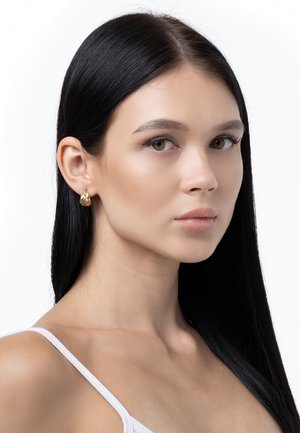 Orecchini d'oro con un design texturizzato, caratterizzati da una forma curva che aggiunge profondità. Indossati da un modello con lunghi capelli scuri e trucco neutro.