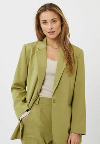 Blazer verde oliva con cierre de un solo botón, solapas de muesca y bolsillos laterales, combinado con una blusa de punto blanco roto. Textura de tela suave.