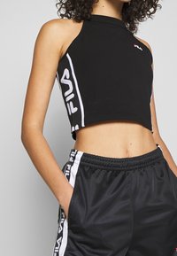 Top corto sin mangas negro con cuello alto, que presenta un logo de FILA blanco impreso en el lateral. Combinado con pantalones de malla negra con rayas laterales.