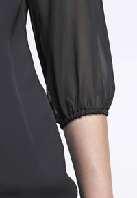 Svart genomskinlig puffärm med elastiskt manschett som bärs över en bar underarm mot en enfärgad ljus bakgrund.