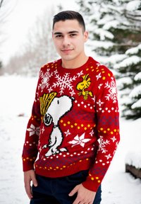 Giovane uomo che indossa un maglione natalizio rosso con Snoopy e Woodstock, in piedi all'aperto in un paesaggio innevato con alberi sempreverdi.