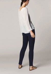 Lichte, loszittende witte trui met 3/4 mouwen, gecombineerd met donkerblauwe slim-fit broek en beige sandalen, gezien vanuit de rug.