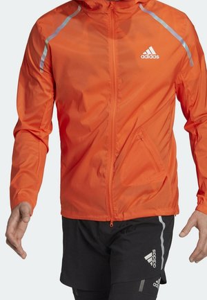 Mężczyzna ubrany w jaskrawopomarańczową kurtkę Adidas zapinaną na zamek z elementami odblaskowymi oraz czarne spodenki Adidas z logo i odblaskowymi paskami.