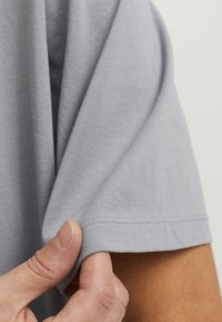 Lichtgrijs katoenen t-shirt met een zachte textuur, met korte mouwen en nette stiksels langs de zoom, gedeeltelijk vastgehouden door een hand.