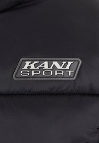 Svart polstret jakke med et rektangulært logo som har teksten "KANI" øverst og "SPORT" under, alt i sølv mot stoffet.