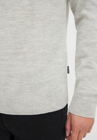 Maglione grigio chiaro lavorato a maglia con finitura testurizzata, polsini a costine e una piccola etichetta del marchio di lato. Indossato sopra pantaloni scuri.