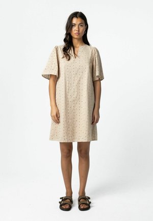 Naine seisab ja kannab beežikat õhukese mustriga lühikest kleiti, millel on lõhkuvad varrukad, ning musti naelutatud sandaale valgel taustal.