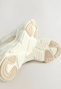 Baskets blanches avec des semelles texturées présentant un motif en vagues et des accents beiges. Le design comprend des panneaux en maille et une semelle rembourrée.