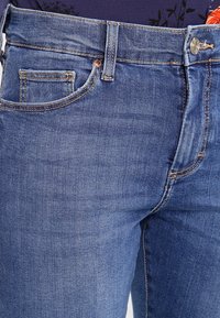 Denimjeans i mellanmörkblått med en figurnära silhuett, utrustade med en framficka, dragkedja och knäppning med metallknapp. Diskreta veckdetaljer.