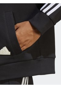 Svart sweatshirt med en framficka och vit tre-randersdesign på ärmarna. Mjuk textur och detaljer med ribbade ärmslut.