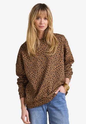 Frau mit langen blonden Haaren, die einen braunen Pullover mit Leopardenmuster, blaue Jeans und goldene Armreifen an einem Handgelenk trägt, steht mit einer Hand in der Tasche.