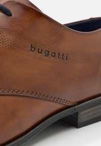 Chaussure en cuir marron présentant une texture lisse, des détails cousus et un logo de marque. Lacets noirs, talon empilé et bout arrondi.