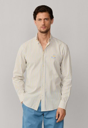 Homme portant une chemise rayée jaune et bleue à manches longues boutonnée avec un logo brodé, associée à un pantalon bleu clair, debout devant un fond uni.