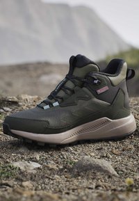 Single green and black hiking boot with rugged sole resting on rocky ground outdoors with blurred mountain background.