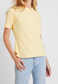 Žluté polo tričko z bavlny s límečkem, krátkými rukávy a pěti knoflíky. V kombinaci s světle modrými džíny.