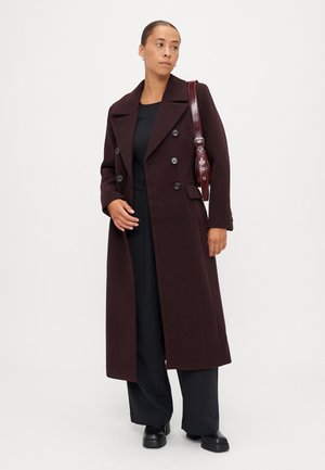Lang bordeaux uldfrakke med dobbeltknap-design, store kraver og sidelommer. Modellen bærer et sort outfit og har en blank bordeaux taske.