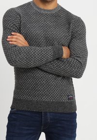 Grå stickad tröja med ett strukturerat mönster, rundad halsringning, ribbade mudd och fåll. Bärs över blå jeans med en logotagg på tröjan.