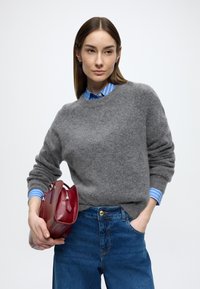 Szürke kötött pulóver, alatta kék-fehér csíkos ing, magasderekú farmernadrág. Piros bőr kézitáskát tart.