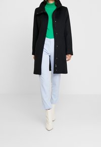 Černý dlouhý kabát s vysokým límcem, vybavený patentkami a zipem. Spárován se zeleným rolákem, světle modrými kalhotami a bílými kotníkovými botami.