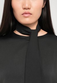 Črna bluza z vezanjem pri vratu, izdelana iz gladkega materiala. Oblikovanje vključuje okroglo ovratnico in minimalistične podrobnosti.