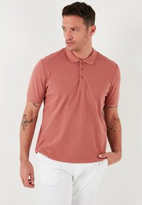 Lühikeste varrukatega polo särk koralltoonis, valmistatud pehmest kangast. Omab klassikalist kraed, kolme nupuga kinnitust ja vabalt langevat lõiget. Valged püksid.