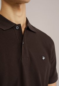 Hnedý polo shirt vyrobený z textúrovaného materiálu, obsahujúci dva gombíky a malé biele vyšívané logo na ľavej hrudi.