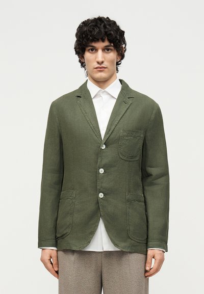 Homme portant un blazer vert olive boutonné avec trois poches, une chemise blanche col à col et un pantalon beige, debout devant un fond uni.