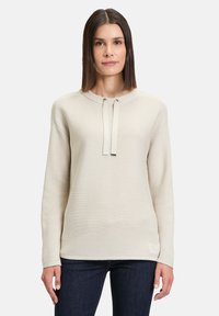 Beige gerippter Strickpullover mit rundem Halsausschnitt, Kordelzug-Details und langen Ärmeln, kombiniert mit dunkelblauen Jeans.