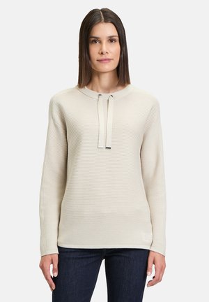 Beige ribbed knit sweater with a rounded neckline, drawstring accents, and long sleeves, paired with dark blue jeans.