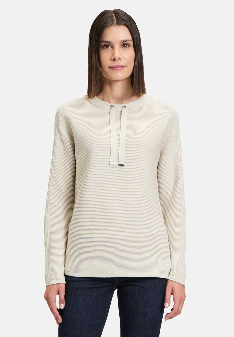 Beige gerippter Strickpullover mit rundem Halsausschnitt, Kordelzug-Details und langen Ärmeln, kombiniert mit dunkelblauen Jeans.