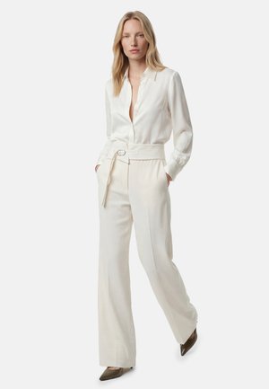 Vrouw draagt een ivoorkleurige zijden blouse met knopen, ingestopt in een hoge taille, wijd gesneden broek met een bijpassende riem en puntige bruine hakken.