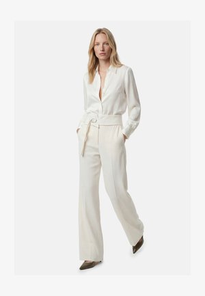 Donna che indossa una camicetta in seta color avorio con bottoni, infilata in pantaloni a vita alta e gamba larga con cintura abbinata e tacchi marroni a punta.