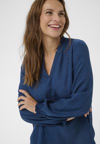 Bluză albastră cu mâneci lungi, decolteu în V și detaliu cu volane la guler. Material moale, texturat, cu manșete elastice, croială ajustată.