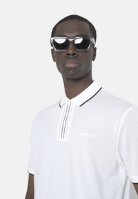 Camisa polo blanca con acentos negros, tapeta de dos botones y logo bordado. El modelo lleva gafas de sol transparentes con un diseño geométrico.