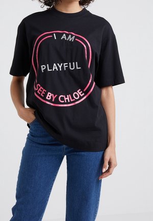 Femme portant un T-shirt noir avec un bord circulaire rose et les textes « I AM PLAYFUL » et « SEE BY CHLOE » associé à un jean bleu.