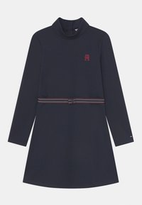 Granatowa sukienka z długim rękawem i wysokim kołnierzem, z czerwonym logo na piersi oraz paskiem w paski w talii. Gładka tkanina, prosty krój.